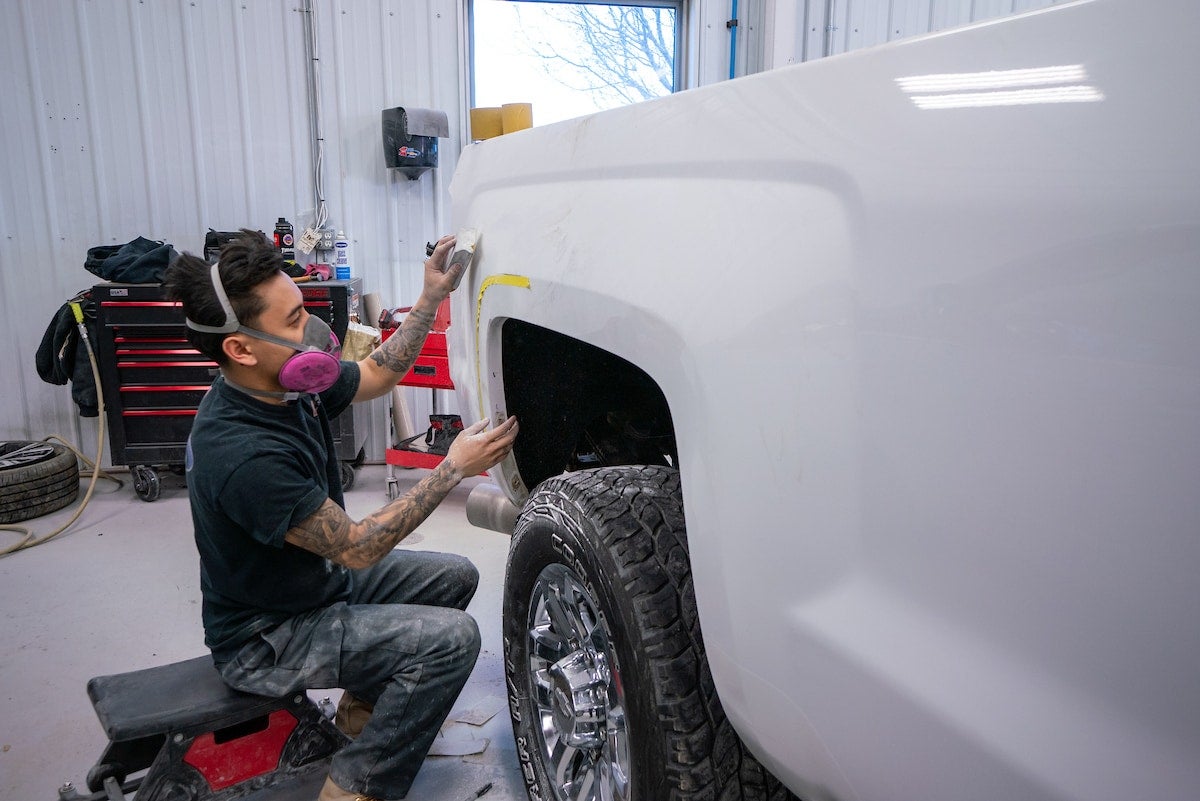 A person wearing a respirator sanding the wheel well of a white pickup truck in a brightly lit garage.